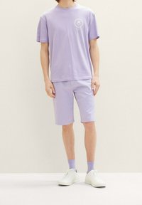 Camiseta de algodón lavanda con mangas cortas y cuello redondo, combinada con pantalones cortos a juego. Zapatillas blancas y calcetines lavanda completan el look.