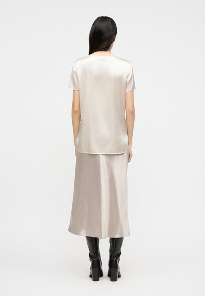 Γυναίκα με μακριά σκούρα μαλλιά που φοράει μια φαρδιά σατέν μπλούζα με κοντά μανίκια και μακρύ φόρεμα που ταιριάζει, συνδυασμένα με μαύρες ψηλές μπότες με τακούνι, γυρισμένη προς τα πίσω.