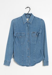Camicia in denim blu chiaro con maniche lunghe, colletto a punta, due tasche anteriori sul petto e chiusura con bottoni. Presenta un orlo curvo e una texture.