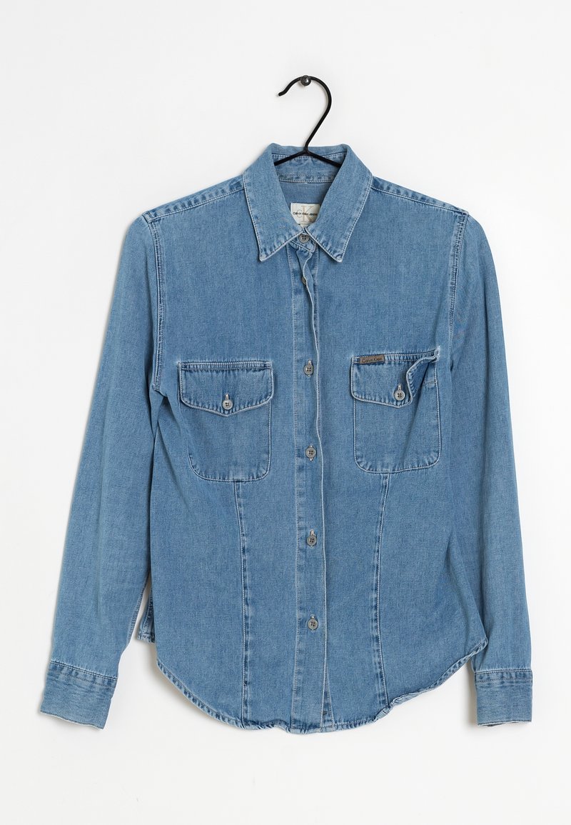 Camicia in denim blu chiaro con maniche lunghe, colletto a punta, due tasche anteriori sul petto e chiusura con bottoni. Presenta un orlo curvo e una texture.
