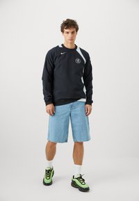 Sort Nike sweatshirt med lysegrå accenter, rundt logo, parret med blå denim shorts og sort-grønne træningssko.