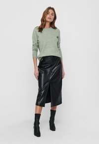Pull en maille vert clair, jupe midi noire en simili cuir avec fente avant, associé à des bottes noires montantes. Design simple et moderne.