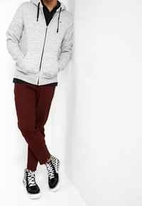 Sudadera gris con cremallera y dos bolsillos, llevada sobre una camiseta negra. Combinada con pantalones granates y zapatillas alta negras con un patrón de tablero de ajedrez.
