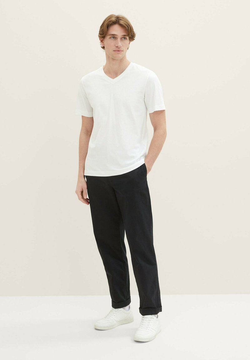 Weißes V-Ausschnitt-T-Shirt kombiniert mit schwarzen Hosen und weißen Sneakers. Glatter Stoff, lockere Passform, kürzere Ärmel und minimalistisches Design.