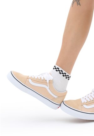 Zapatillas de ante beige con detalles y cordones blancos. Calcetines de tobillo a rayas en blanco y negro visibles. Diseño de corte bajo con suelas de goma.