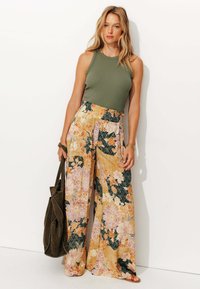 Pantalons larges à motifs floraux orange, rose et vert ; taille élastique. Débardeur vert olive ; portant un sac fourre-tout vert foncé.