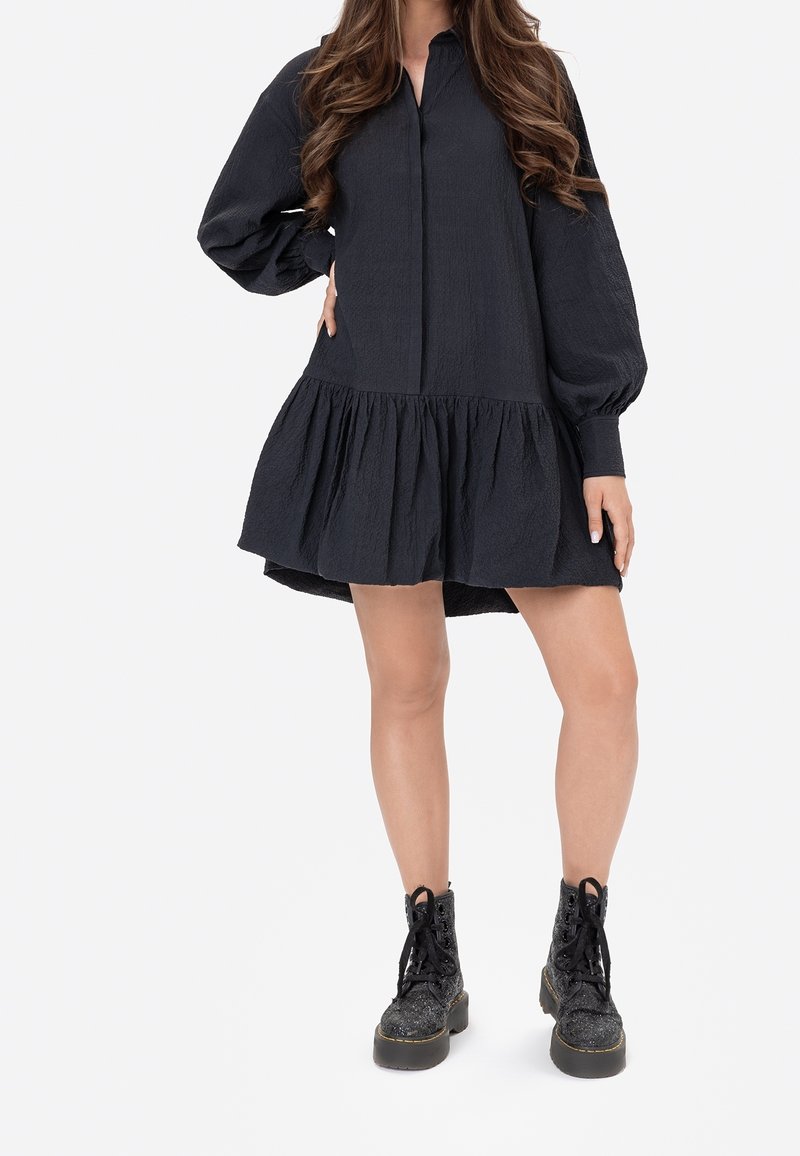 Black, long-sleeve dress with a textured finish, featuring a flared bottom and a collared neckline, paired with black lace-up boots.