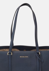 Sac fourre-tout en cuir bleu marine à texture granulée avec deux poignées de épaule, présentant un détail de logo doré et un intérieur spacieux. Texture lisse et design robuste.