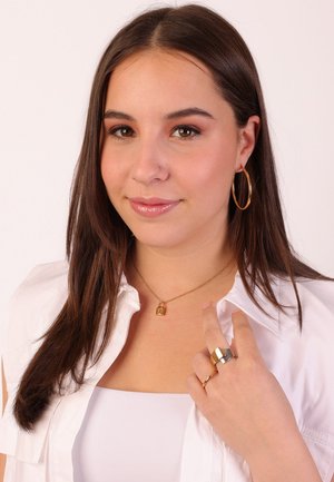 Gouden ketting met hangslot hanger, grote gouden creolen, zilveren en gouden ringen, gedragen met een witte blouse en een neutrale achtergrond.