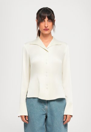 Young woman with dark hair wears an off-white buttoned blouse and light blue jeans, standing against a plain white background.