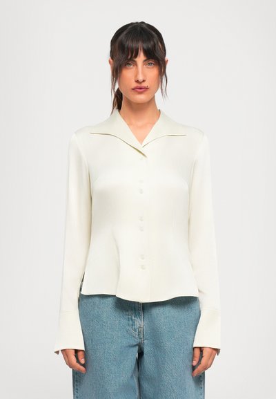 Jonge vrouw met donker haar draagt een lichtbeige blouse met knopen en een lichte spijkerbroek, staand tegen een witte achtergrond.