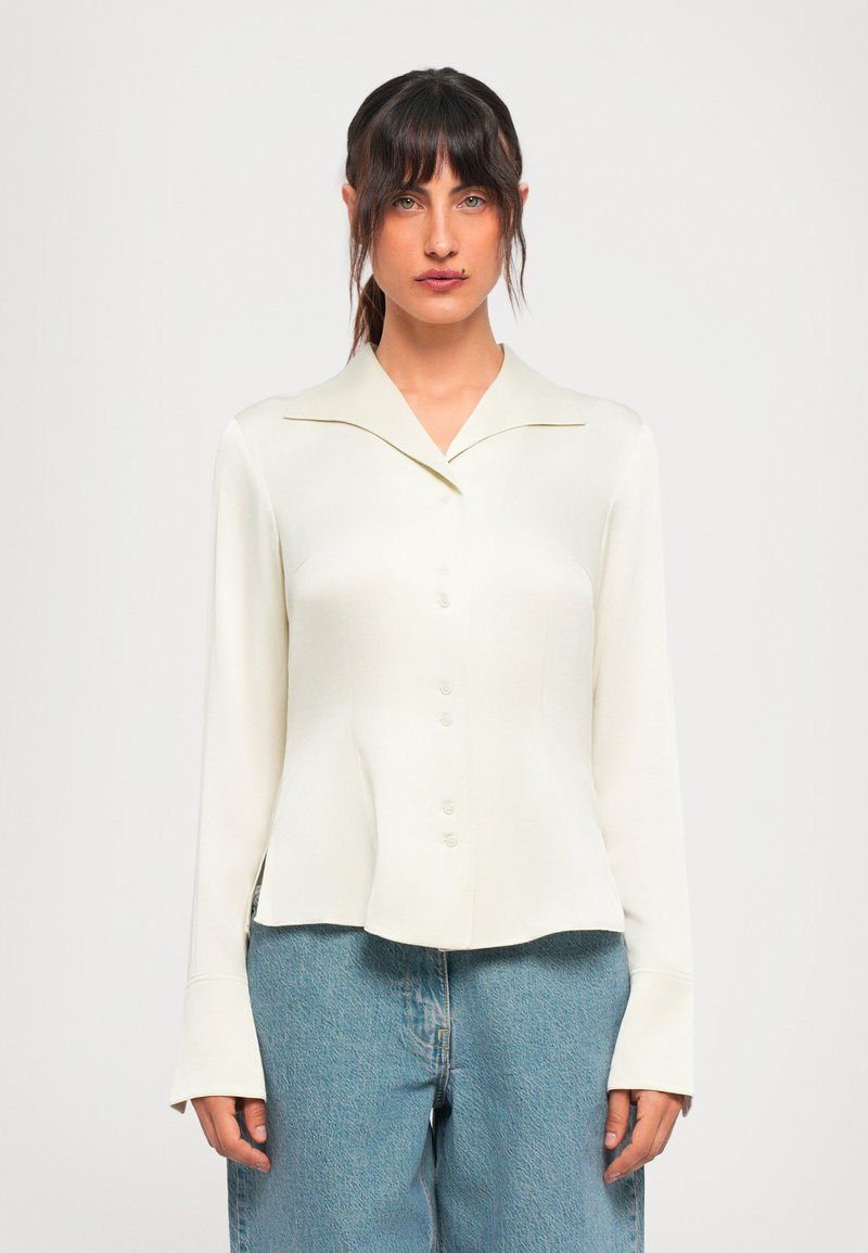 Young woman with dark hair wears an off-white buttoned blouse and light blue jeans, standing against a plain white background.