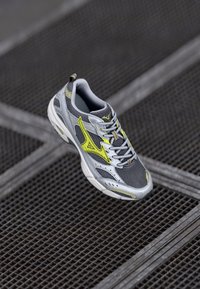 Chaussure de sport grise avec tige en maille, accents jaunes et fermeture à lacets. Semelle texturée, éléments réfléchissants et col rembourré pour le confort.