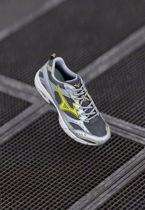 Chaussure de sport grise avec tige en maille, accents jaunes et fermeture à lacets. Semelle texturée, éléments réfléchissants et col rembourré pour le confort.