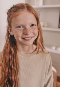 Una ragazza con lunghi capelli rossi ondulati e lentiggini indossa un maglione beige a coste, sorridendo. Lo sfondo è dolcemente sfocato con colori chiari.