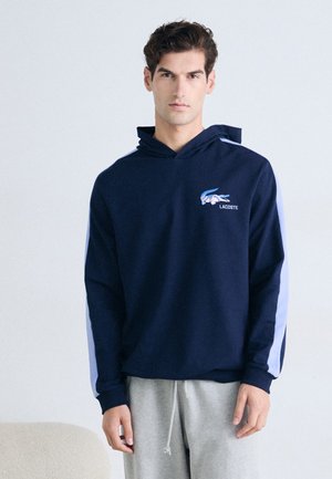Hanorac cu glugă de culoare bleu marin, cu logo Lacoste; dispune de dungi laterale de culoare bleu deschis și o buză în formă de cangur; fabricat dintr-un material moale și texturat.