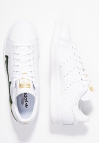 Valkoiset tennarit, joissa on teksturoidut vihreät ja kultaiset korostukset sivulla, kultaiset brändäykset, perinteiset nauhat ja rei'itetyt yksityiskohdat.