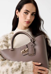 Mujer con cabello oscuro que lleva pendientes de oro y un suéter color crema, sostiene un bolso taupe de Guess con herrajes dorados cerca de su pecho.