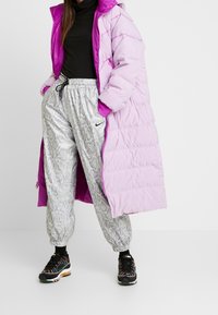 Lång pufferjacka i ljuslila med en kontrasterande magenta insida, kombinerad med silverfärgade byxor i ormtryck och svarta sneakers med färgglada accenter.