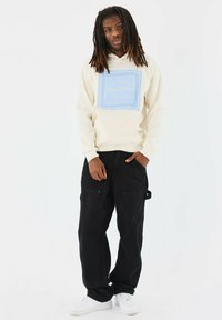Felpa beige con un grafico quadrato blu con testo; abbinata a pantaloni cargo neri e sneakers bianche; fit casual, stile rilassato.