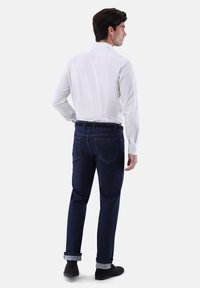 Ein weiß-blau gestreiftes Langarmhemd, kombiniert mit dunkelblauen Jeans, die umgekrempelte Bündchen und ein glattes Finish aufweisen. Schwarze Schuhe runden das Outfit ab.