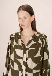 Femme avec des cheveux bruns raides portant un chemisier à boutons avec des motifs de feuilles abstraites verts et beiges sur un fond clair uni.