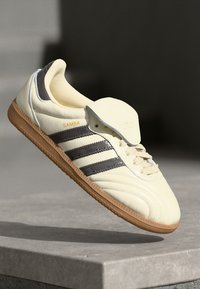 Crème leren sneaker met drie donkere strepen, rubberen gumzool, gevoerde kraag en "SAMBA" logo op de tong. Voorzien van textuur aan de zijkanten.