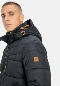 Marineblaue Steppjacke mit einer gesteppten Optik, abnehmbarer Kapuze, Lederapplikationen und einem Reißverschluss vorne. Innenfutter aus Fleece.