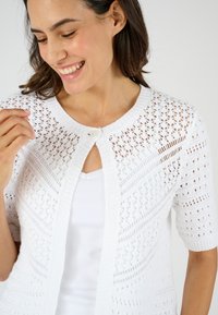 Witte gebreide cardigan met een decoratief opengewerkt patroon, korte mouwen en een knoopsluiting bij de hals.