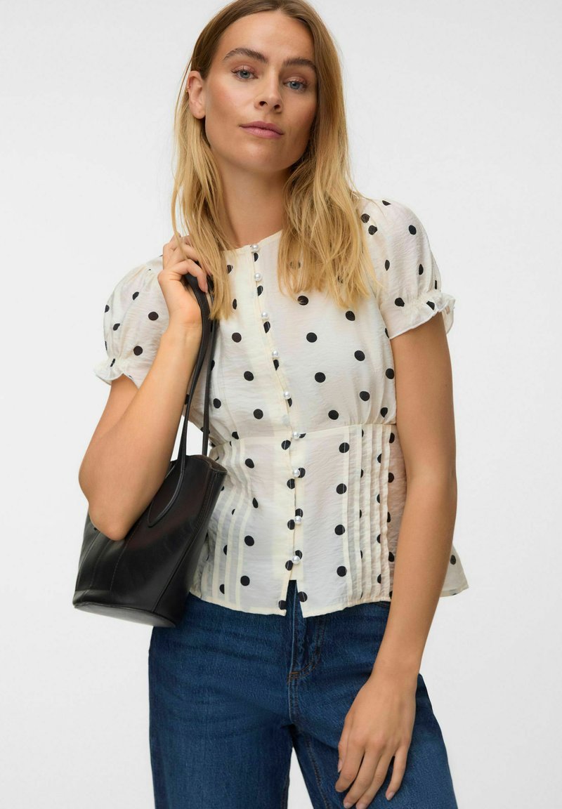 Femme aux cheveux blonds portant un chemisier blanc à pois et un jean bleu, tenant un sac à main noir sur l'épaule devant un fond uni.