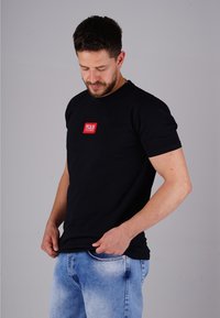 Mann mit Bart, der den Saum eines schwarzen T-Shirts mit rotem Logo anpasst, trägt hellblaue Jeans und steht vor einem schlichten grauen Hintergrund.