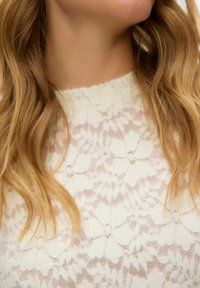 Parte superior de encaje con un delicado patrón floral en color blanco hueso. La tela es semitransparente con un acabado texturizado y un escote alto.