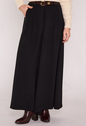 Donna con pantaloni neri a gamba larga, cintura marrone, maglione beige e stivaletti con tacco marrone scuro, in piedi con una mano in tasca.