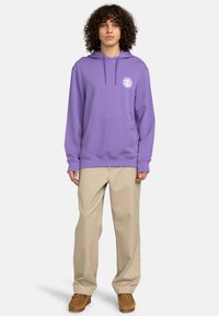 Purple hoodie made of cotton, featuring a front pocket and drawstring hood. Worn with tan trousers and light brown footwear.
