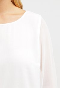 Blusa blanca con escote redondo y mangas largas y transparentes. La tela tiene un acabado ligeramente texturizado, creando una apariencia ligera y etérea.
