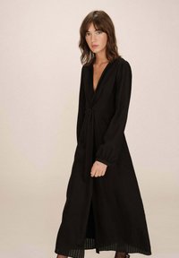 Robe noire à manches longues avec un décolleté en V profond et une taille cintrée. Le tissu est texturé, avec un ourlet plissé pour plus de détail.