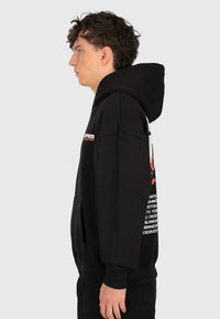 Un jeune homme en sweat à capuche noir avec un texte rouge et blanc se tient de profil contre un fond clair.