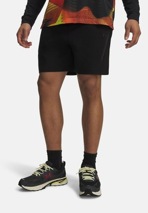 Man die zwarte sportshorts, zwarte sokken en zwarte sneakers met gele en rode details draagt, staand tegen een witte achtergrond.