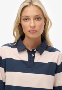 Blonde vrouw met blauwe ogen in een marineblauw en wit horizontaal gestreept poloshirt, kijkt recht vooruit met een neutrale uitdrukking.