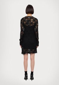 Abito in pizzo nero con maniche lunghe, intricati motivi floreali e orlo scallop. Lunghezza tunica, con un design trasparente e una silhouette aderente.