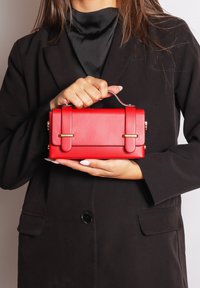 Borsa rettangolare rossa con una texture in pelle liscia, dettagli in metallo dorato e un manico corto, tenuta da una persona che indossa un blazer nero.