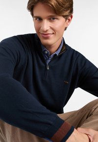 Marineblauer Pullover mit halbem Reißverschluss und gerippten Bündchen, ausgestattet mit einem kleinen gestickten Logo auf der Brust und einem hellblauen Hemd darunter.