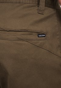 Barna szövet cargo nadrág, sima textúrával, hátsó zsebbel és az oldalán a Volcom logójával ellátott címkével.