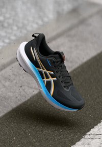 Schwarzer Sportschuh mit strukturiertem Obermaterial, ausgestattet mit weißen und goldenen Akzenten sowie einer blauen Sohle. Leichtes Design mit Schnürsystem.