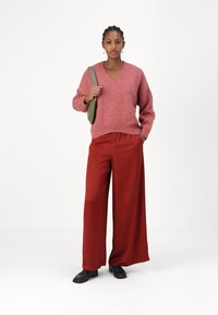 Roze v-halstrui met een pluizige textuur gecombineerd met brede rode broeken. Accessoires zijn een groene schoudertas en zwarte schoenen.