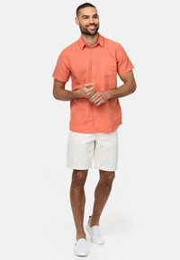 Korte ærmer, koralfarvet linnedskjorte med brystlomme, parret med hvide denimshorts og hvide slip-on sko; glat tekstur og afslappet pasform.