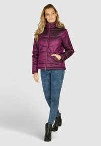 Paarse pufferjack met een hoge kraag, horizontaal gestikt ontwerp, ritssluiting en zijzakken. Gecombineerd met blauwe jeans en zwarte laarzen.