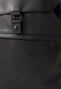 Black leather backpack featuring a magnetic snap closure, front zipper pocket, and a brand tag. Smooth texture with stitched detailing.