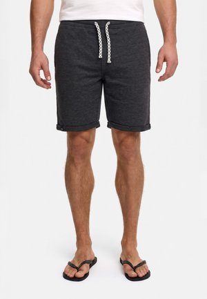 Uomo che indossa pantaloncini grigio scuro con risvolti arrotolati, cordino bianco, camicia bianca e infradito nere, in piedi su uno sfondo bianco.
