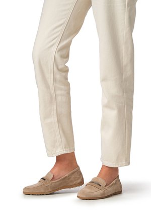 Mocasines de ante beige con suela plana, con un corte sencillo y un adorno negro decorativo en la parte superior. Usados con pantalones de color claro.
